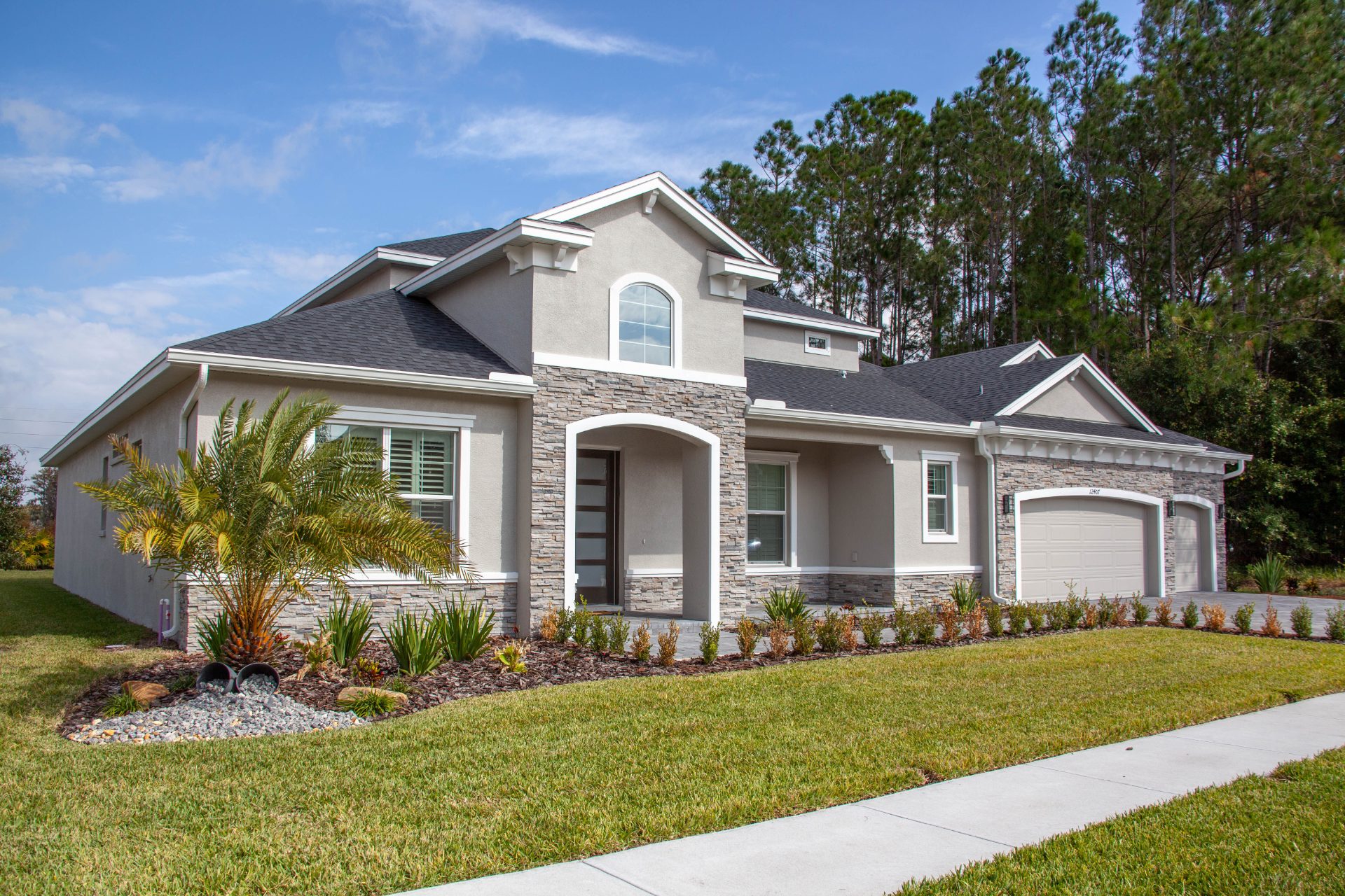 New single-story stucco home with stone accents and a three-car garage, backed by tall pines
